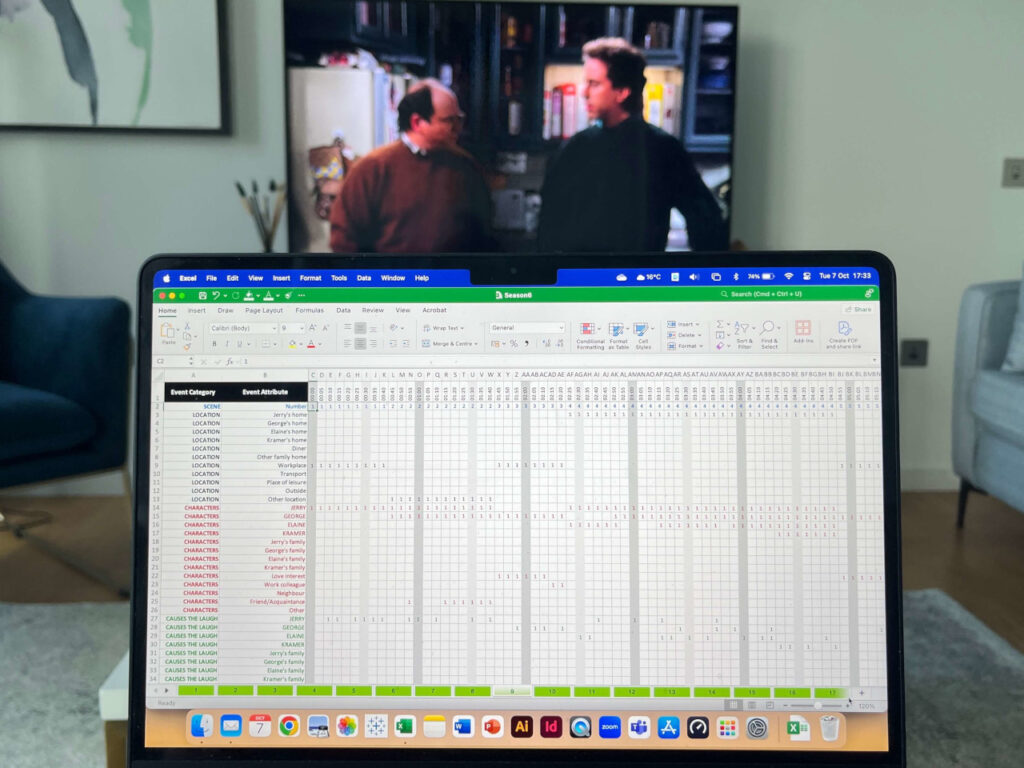 Im Vordergrund steht ein aufgeklappter Laptop, auf dessen Bildschirm eine Tabellenkalkulation zu sehen ist. Im Hintergrund steht ein Fernseher, auf der eine Seinfeld-Folge läuft.
